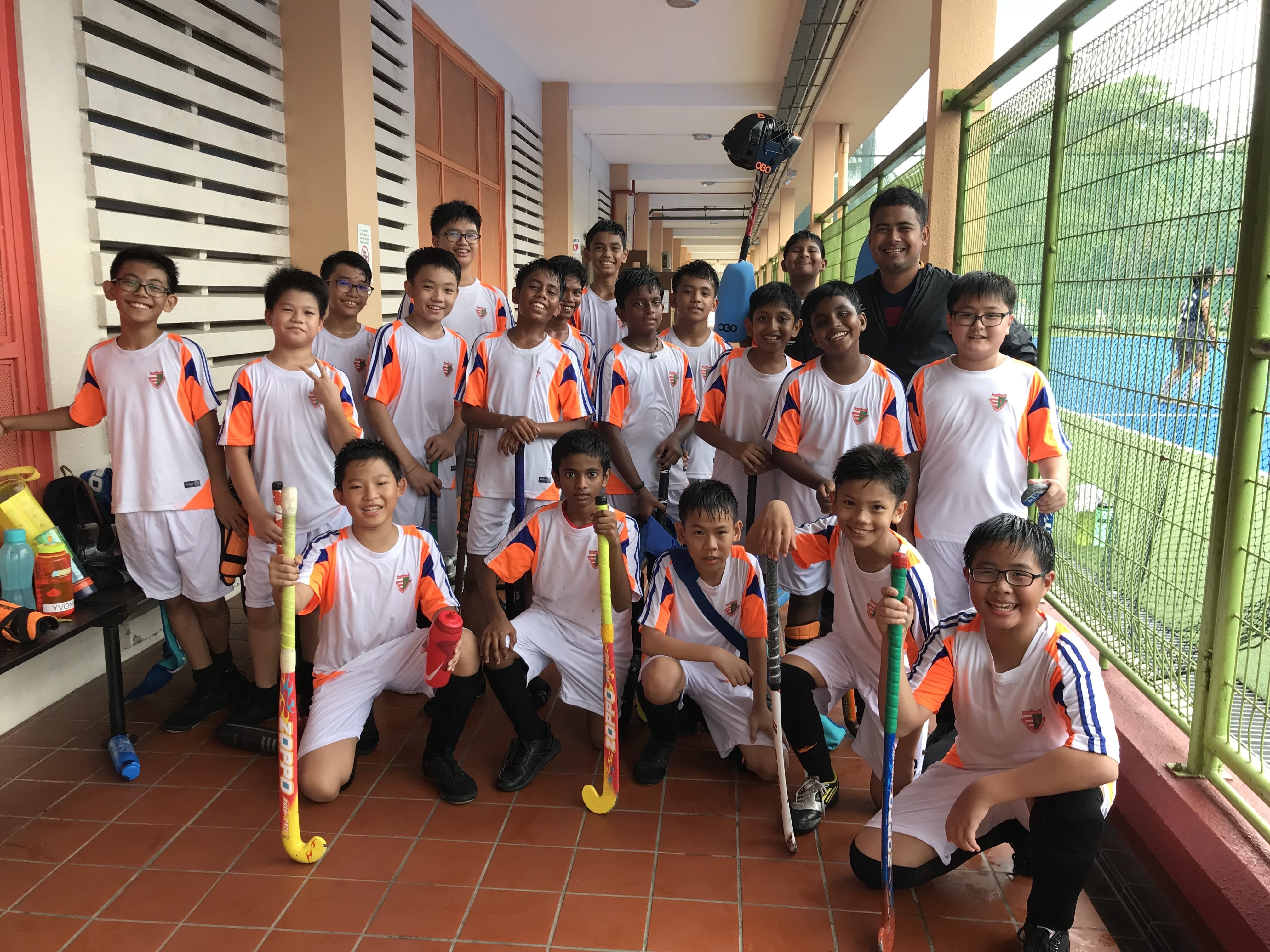 A youth field hockey team in white and orange uniforms poses with sticks in a school corridor with an adult coach.